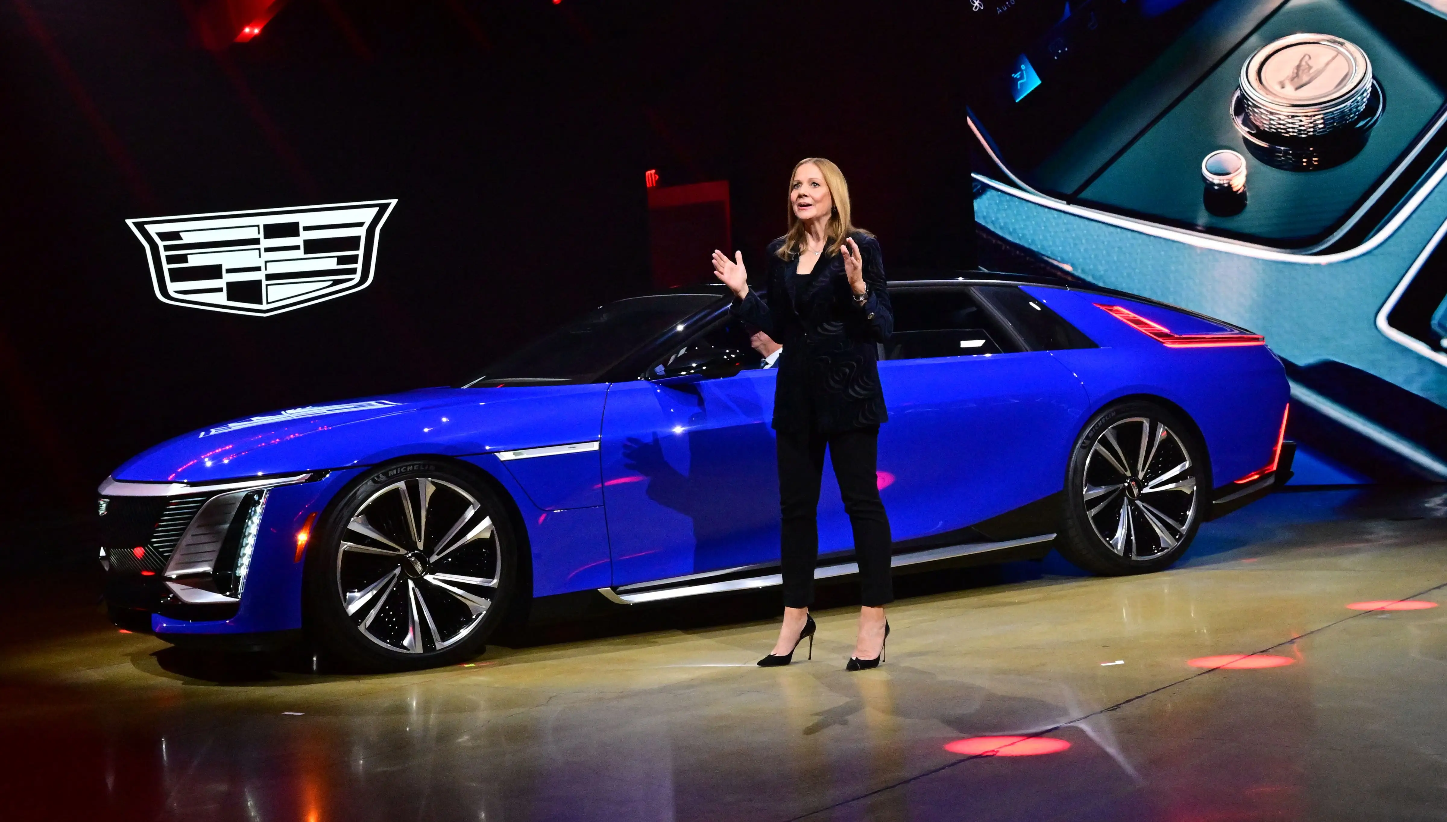 GM CEO Mary Barra, dressed in a black suit, stands on stage next to a bright blue Cadillac Celestiq. The car is a large luxury electric sedan.