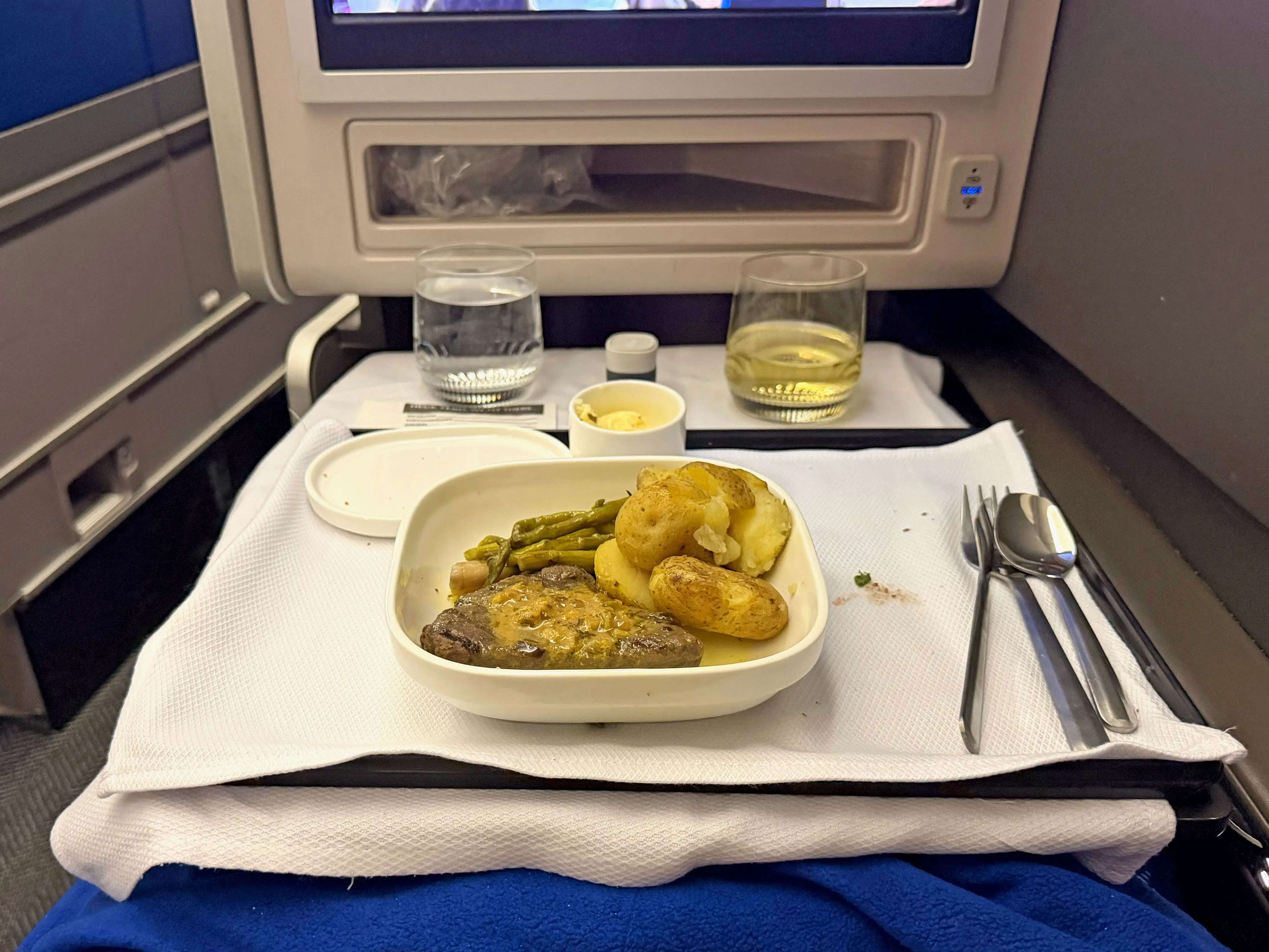 Steak, potatoes, and vegetables on a plate, along with silverware, water, and a glass of white wine on a plane tray table.