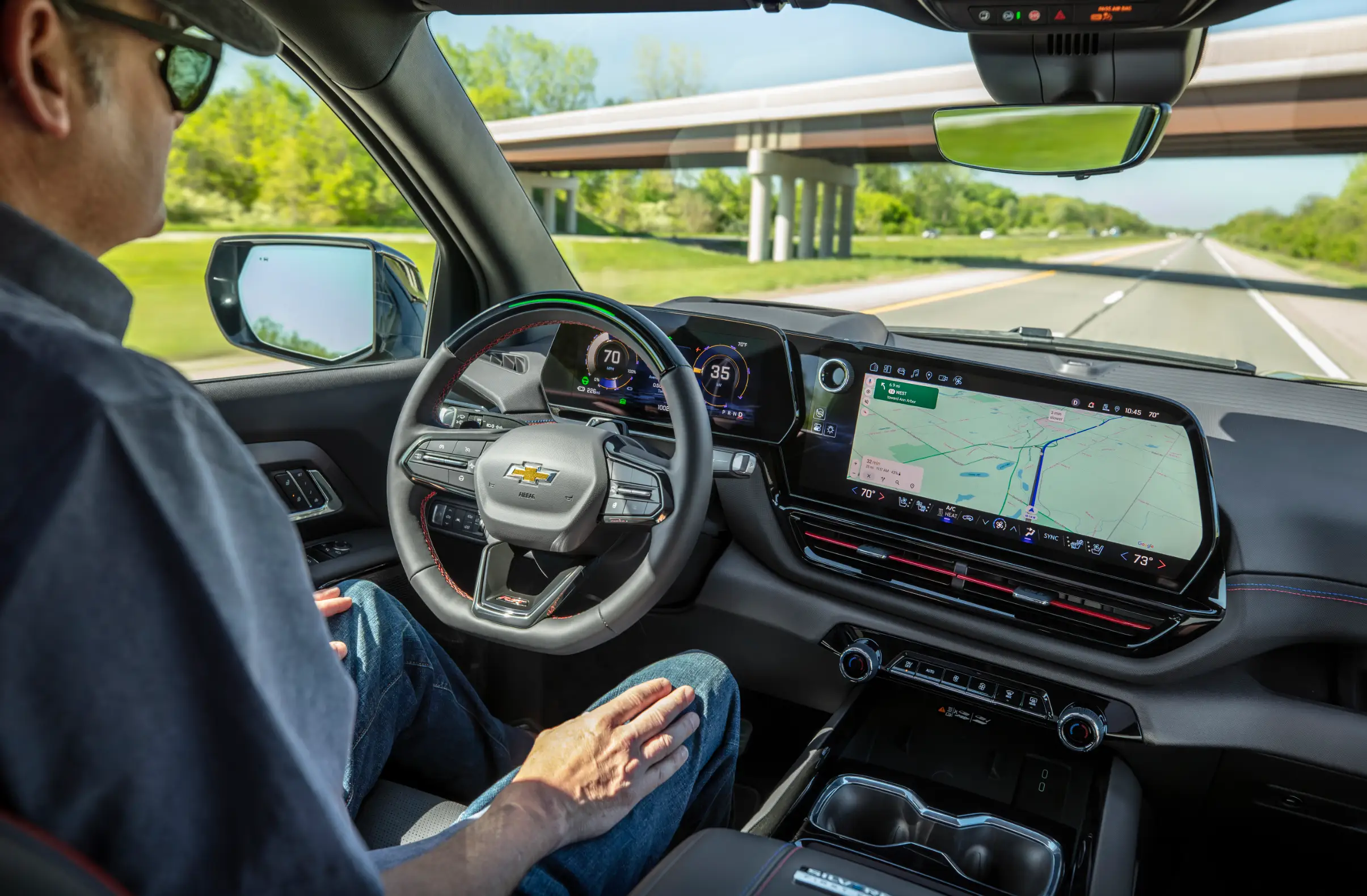 A man in blue jeans is driving down a two-lane highway under an overpass. The driver looks forward and doesn't have his hands on the wheel.