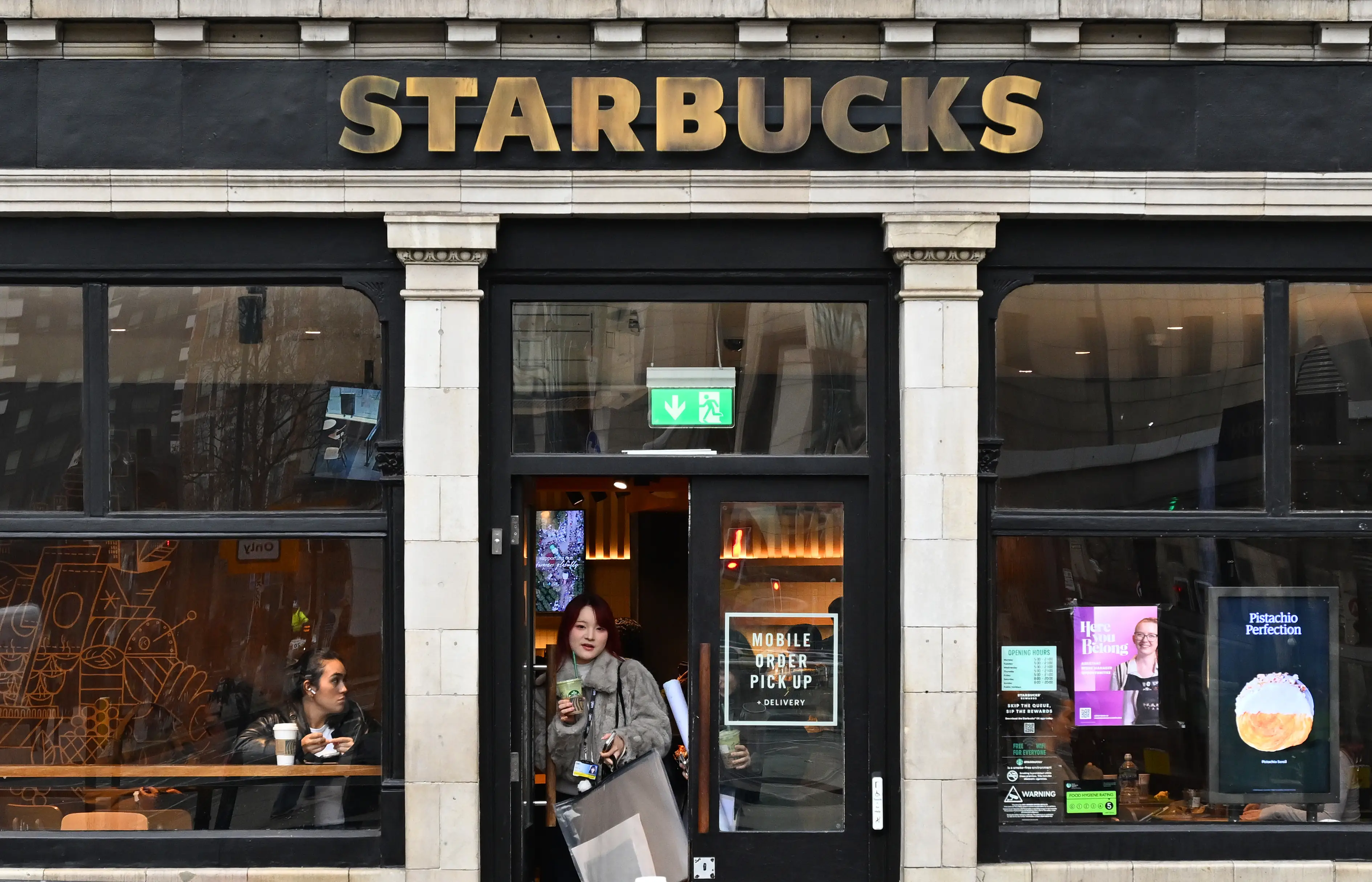 The storefront of a Starbucks café with a customer exiting, drink in hand.