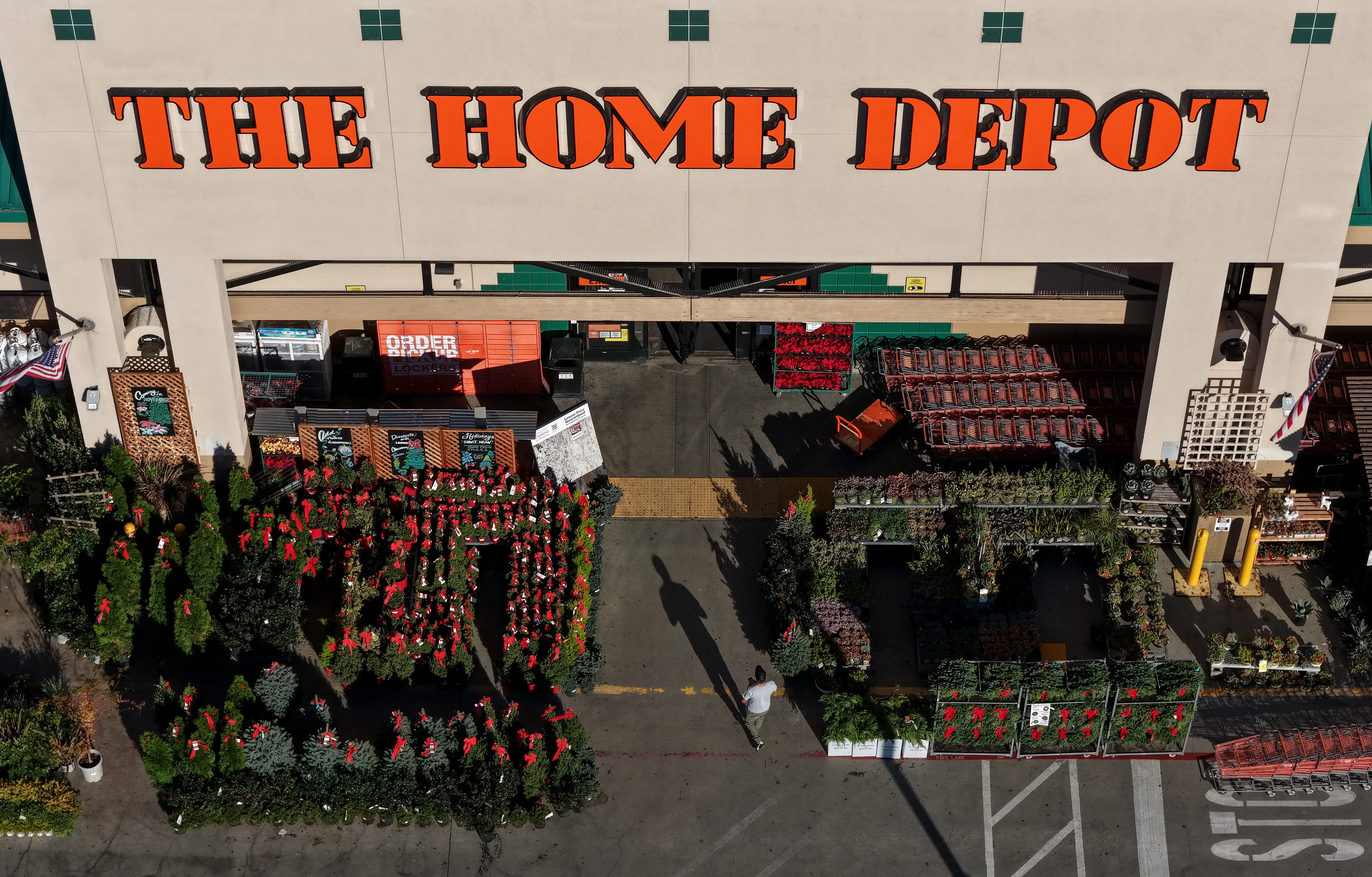 The entrance to a Home Depot store, with a customer walking into the store and a selection of plants outside of it.