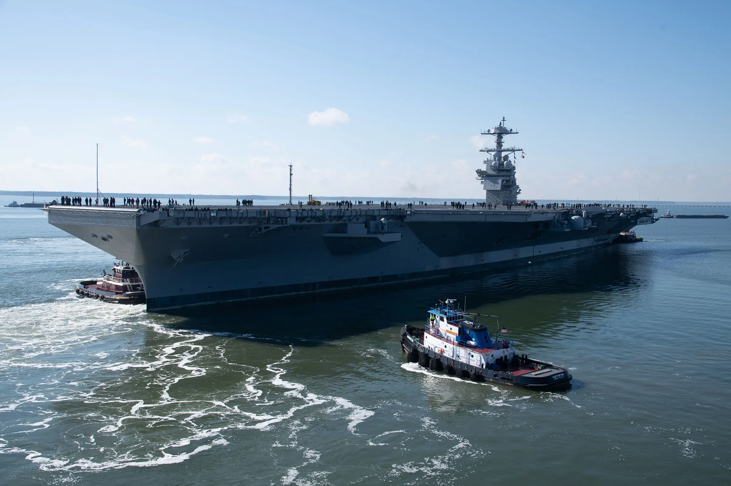 The US Navy USS John F. Kennedy aircraft carrier sitting in blue water with a lightly cloudy sky in the background.