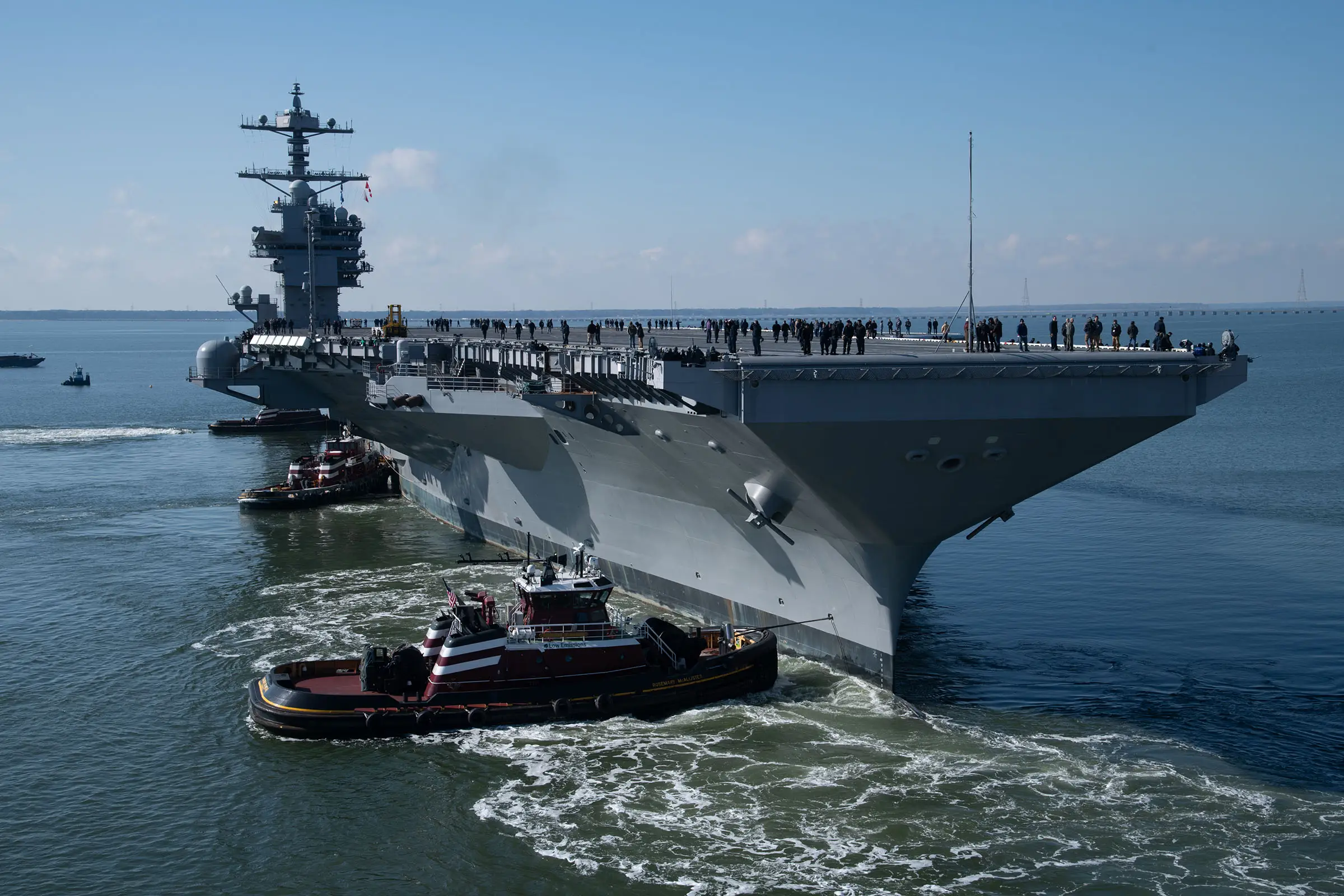 The US Navy USS John F. Kennedy aircraft carrier sitting in blue water with a lightly cloudy sky in the background. Two smaller boats are sailing next to it.