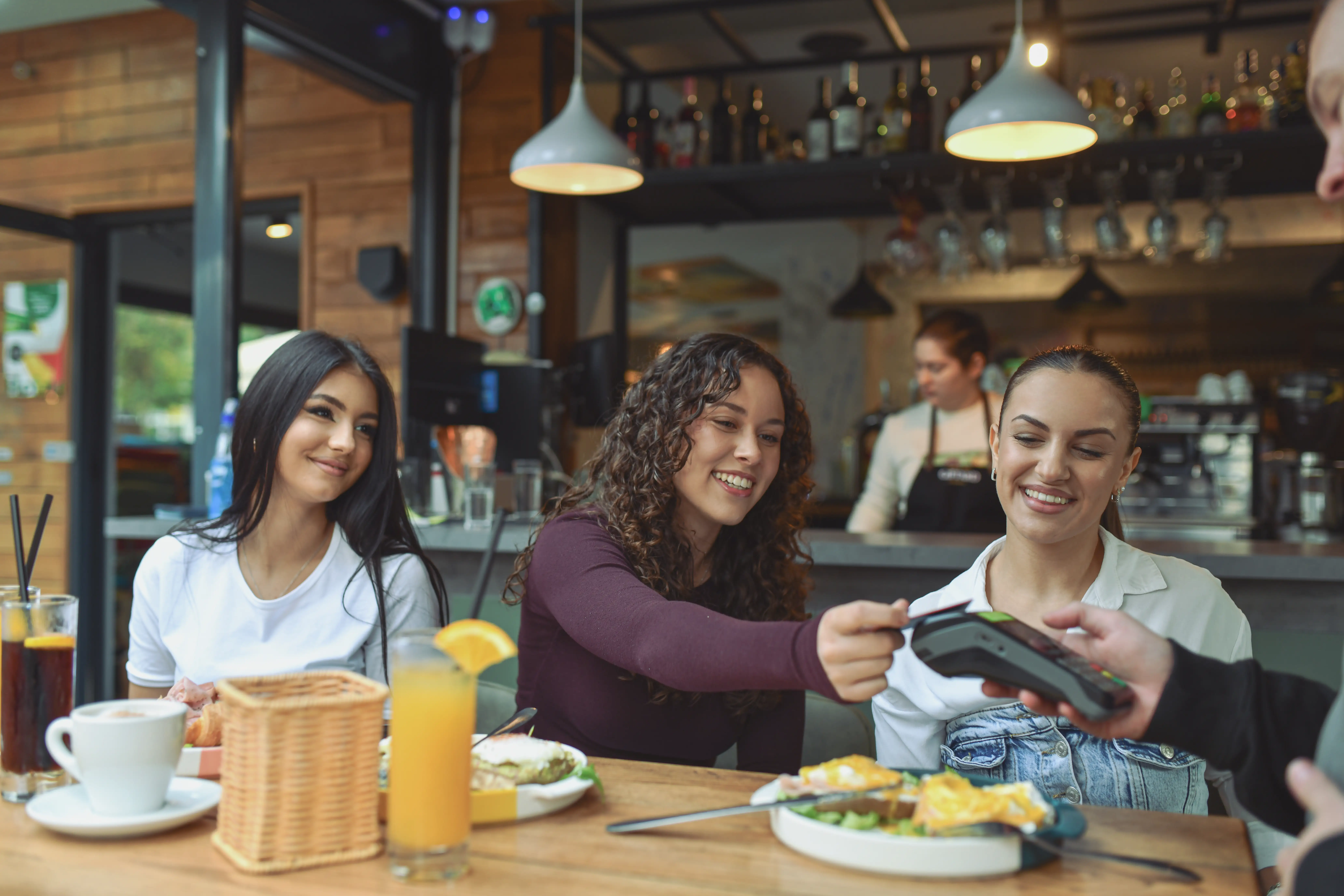 Three girls seated at a resturant, the middle one is tapping her credit card to pay.
