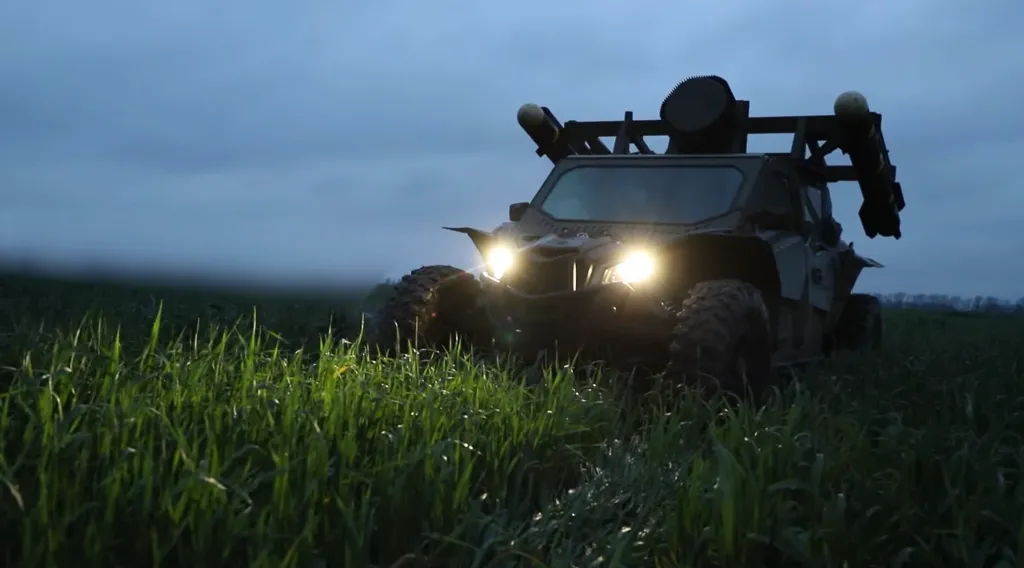 A vehicle-mounted missile launcher with its headlights on, parked in a field of tall green grass under a twilight sky.