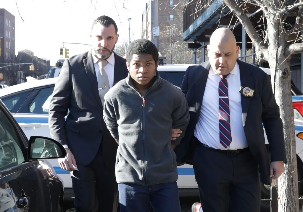 De'Ovryion Elijha Ray, in a gray jacket, is escorted by two men in suits, with police cars in the background.