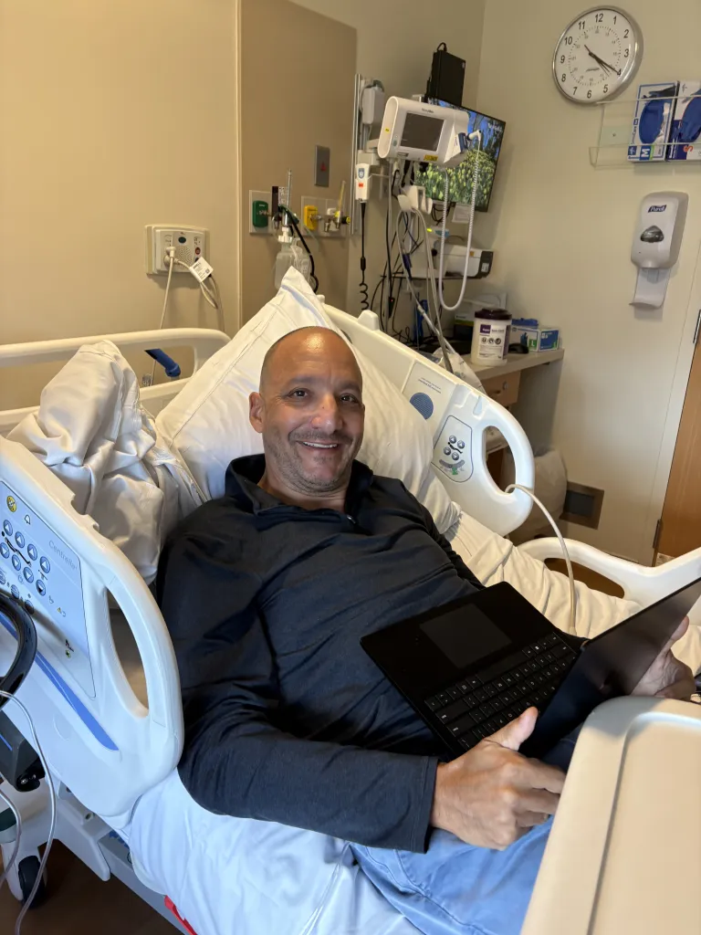 A man smiling from a hospital bed while using a laptop.