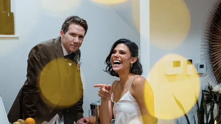 A man in a brown suit looks at the camera while a woman in a white dress laughs and points.