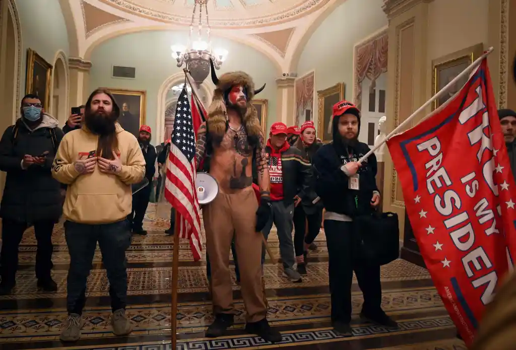 Man in fur hat with horns and face paint carries an American flag inside a building, surrounded by other people.