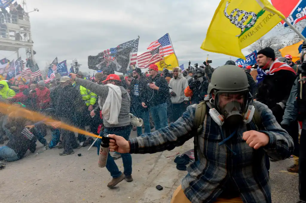 A man in a gas mask and military helmet sprays an orange substance from a can into a crowd of protestors under a cloudy sky.