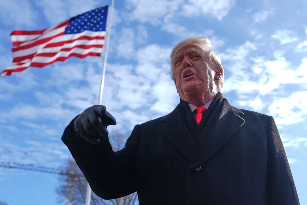 Donald Trump in a black coat and red tie, speaking with an American flag and blue sky in the background.