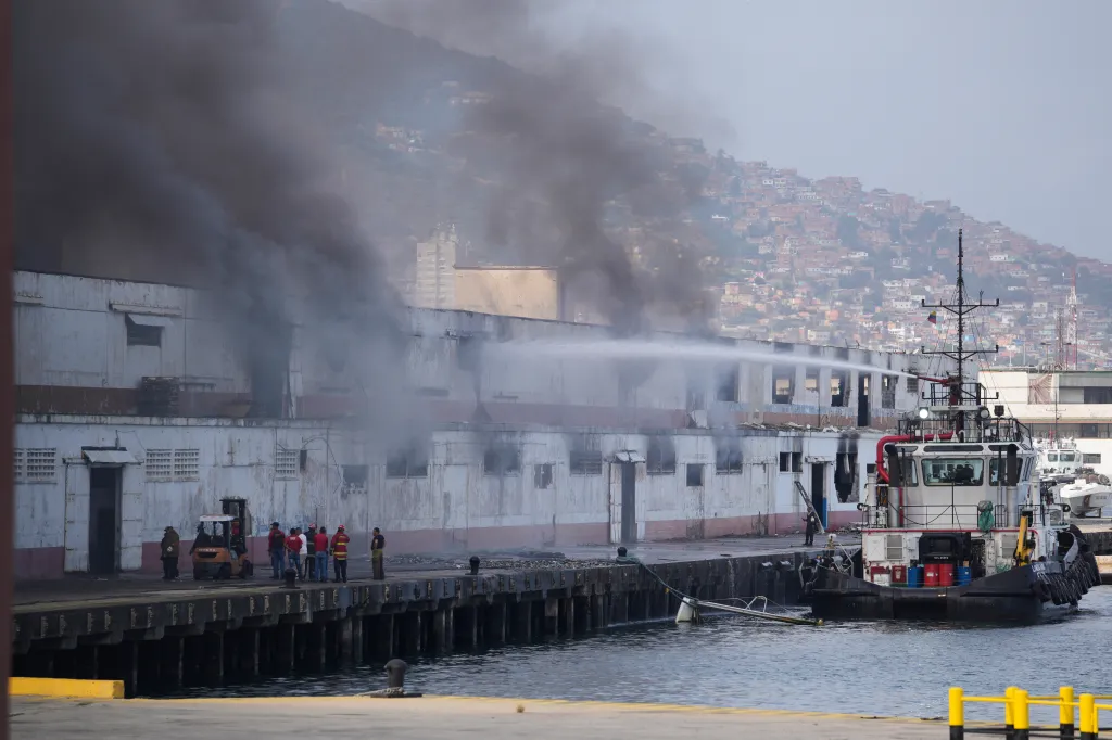 A burning building on a dock with a fireboat spraying water, and a mountain with houses in the background.