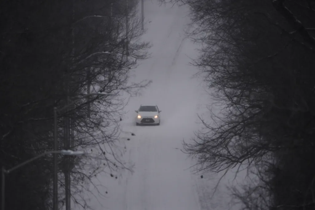 A white car drives down a snowy road, framed by dark, bare trees.