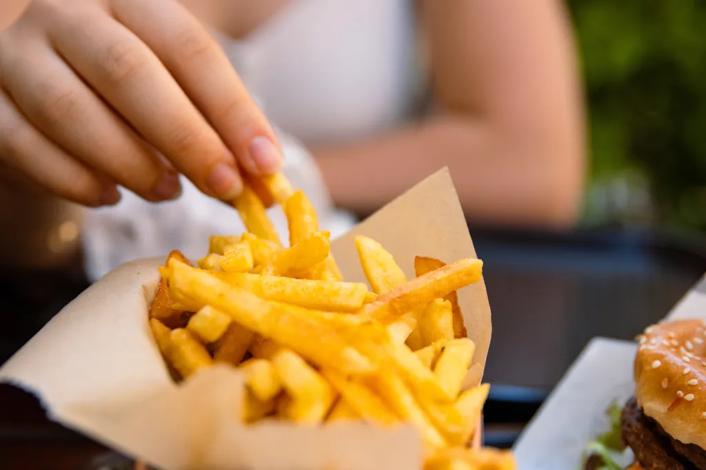 A person's hand reaching for a crispy golden fry from a container, with a burger partially visible.