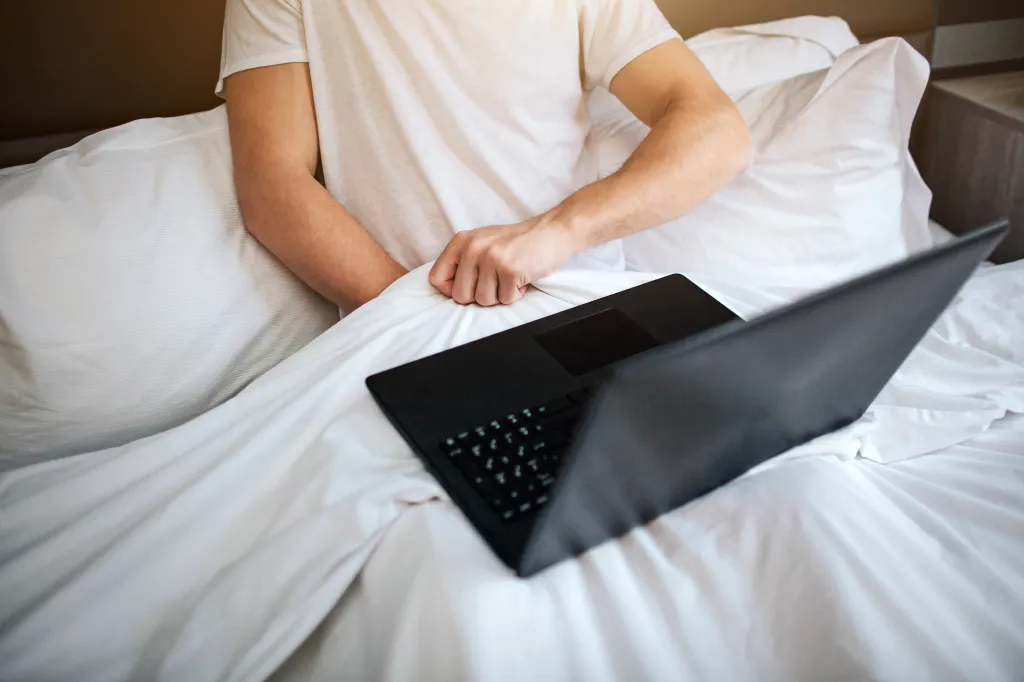 Young man sit in bed early morning. He hold hand under white blanket and masturbating. Laptop on his legs. Watching video.