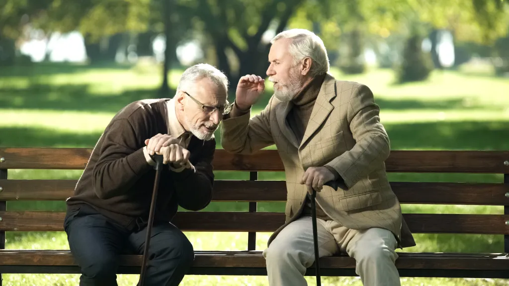Two elderly men sitting on a park bench; one leans in to speak to the other who cups his ear, indicating hearing impairment.
