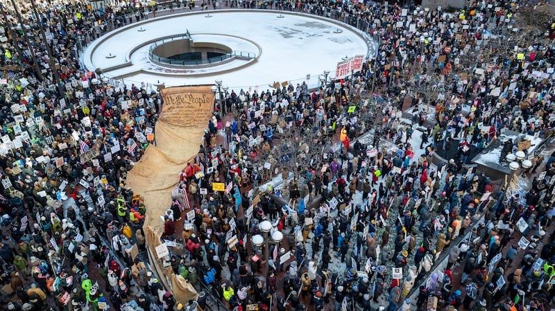 A crowd of protestors surround a draped banner depicting the opening preamble of the U.S. constitution "We The People"