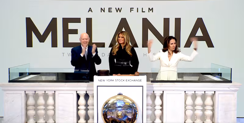 A woman gesticulates while standing beside Melania Trump in front of a sign saying "Melania: A New Film."