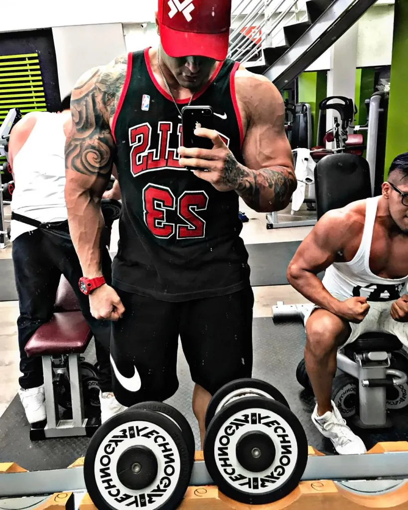 A bodybuilder in a black Bulls jersey and red cap taking a mirror selfie in a gym.