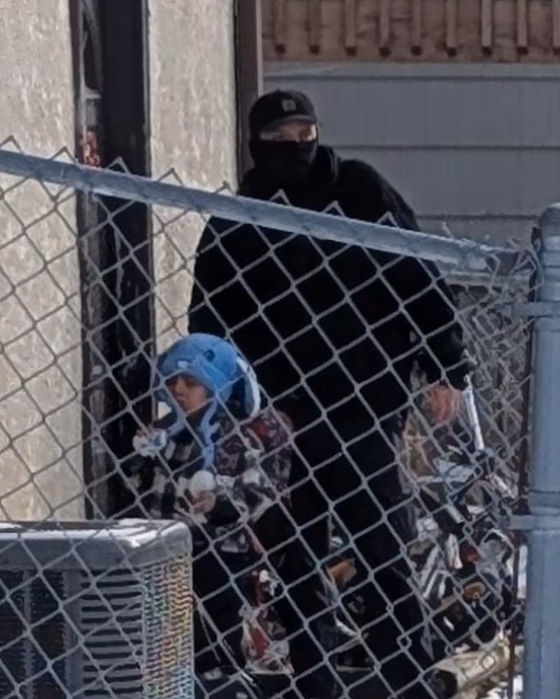 A masked agent ushers the child through a yard.