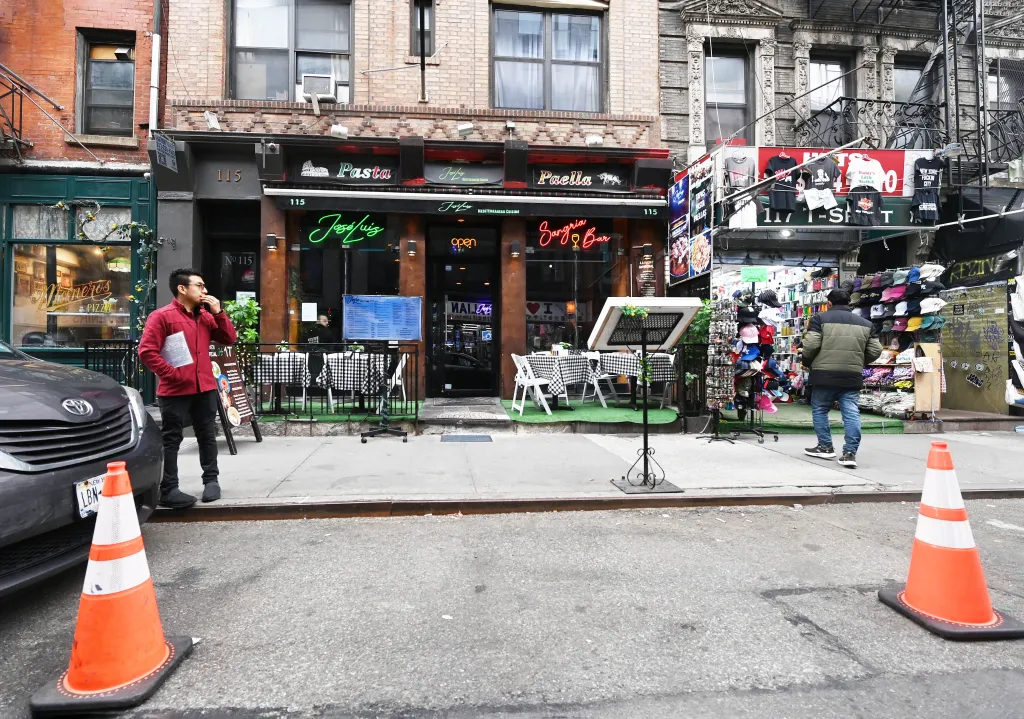 A man in a red jacket stands beside a car parked on the street with the “Jose Luis” restaurant, featuring outdoor seating, in the background.