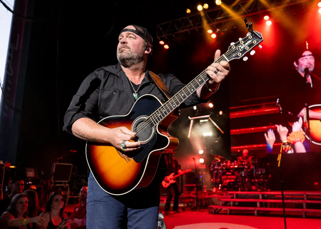 A man wearing a black shirt, a black cap, and jeans playing an acoustic guitar on a stage with red and yellow lights.