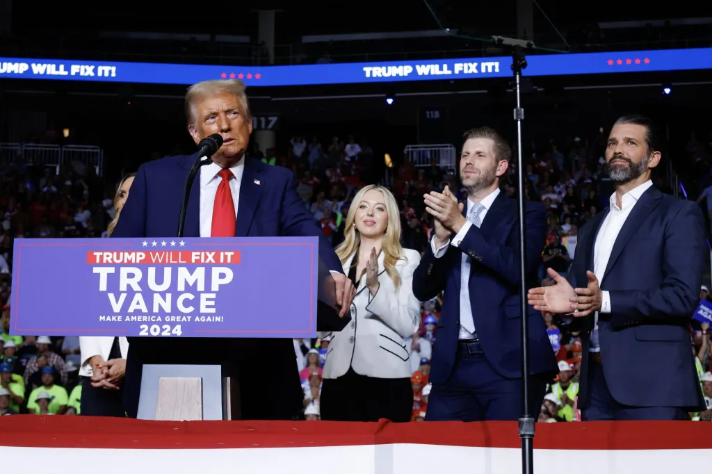 Donald Trump speaking at a rally with Eric Trump, Donald Trump Jr., and Tiffany Trump, in front of a sign that reads