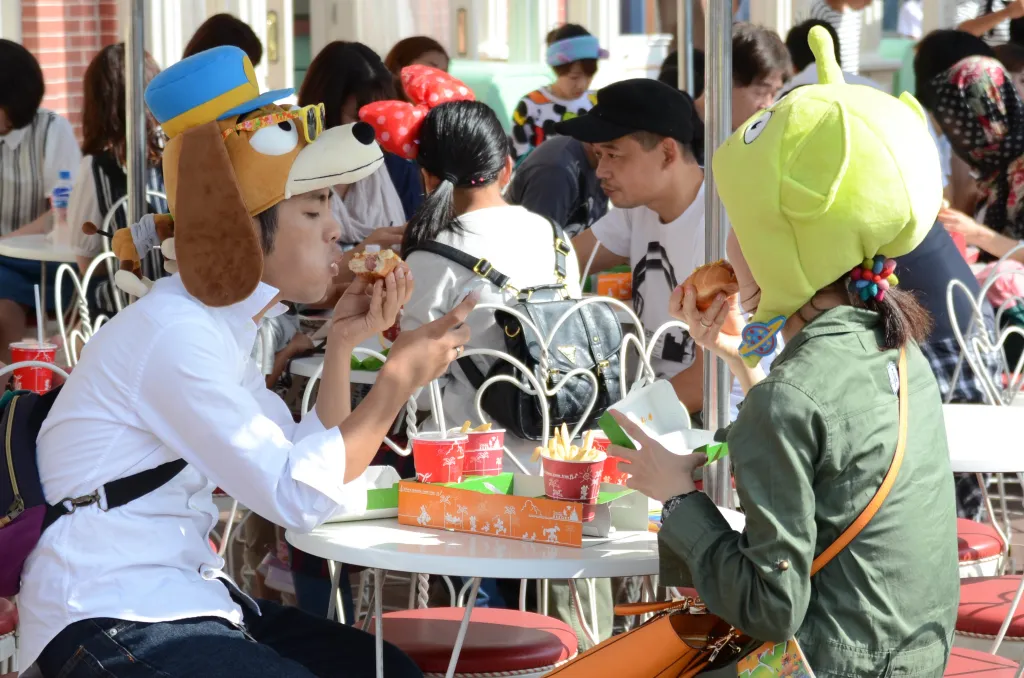 Two people wearing character hats eat hot dogs and fries at an outdoor cafe.