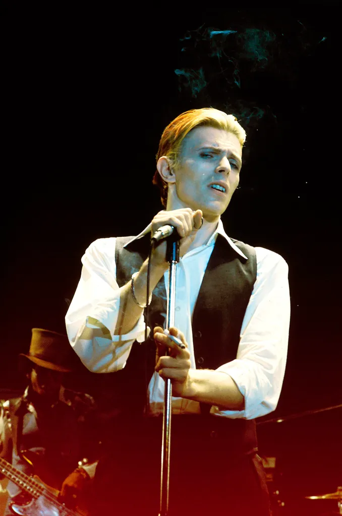 David Bowie performing on stage, holding a microphone in one hand and a cigarette in the other.