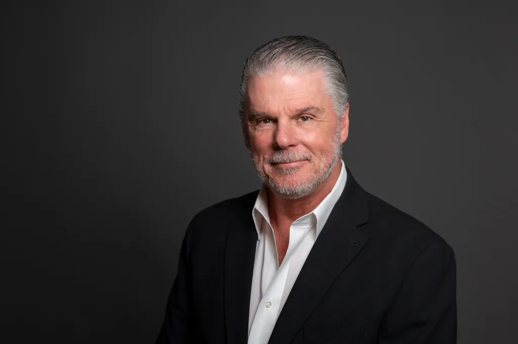 Headshot of a man with gray hair and a beard, wearing a black suit and white shirt.