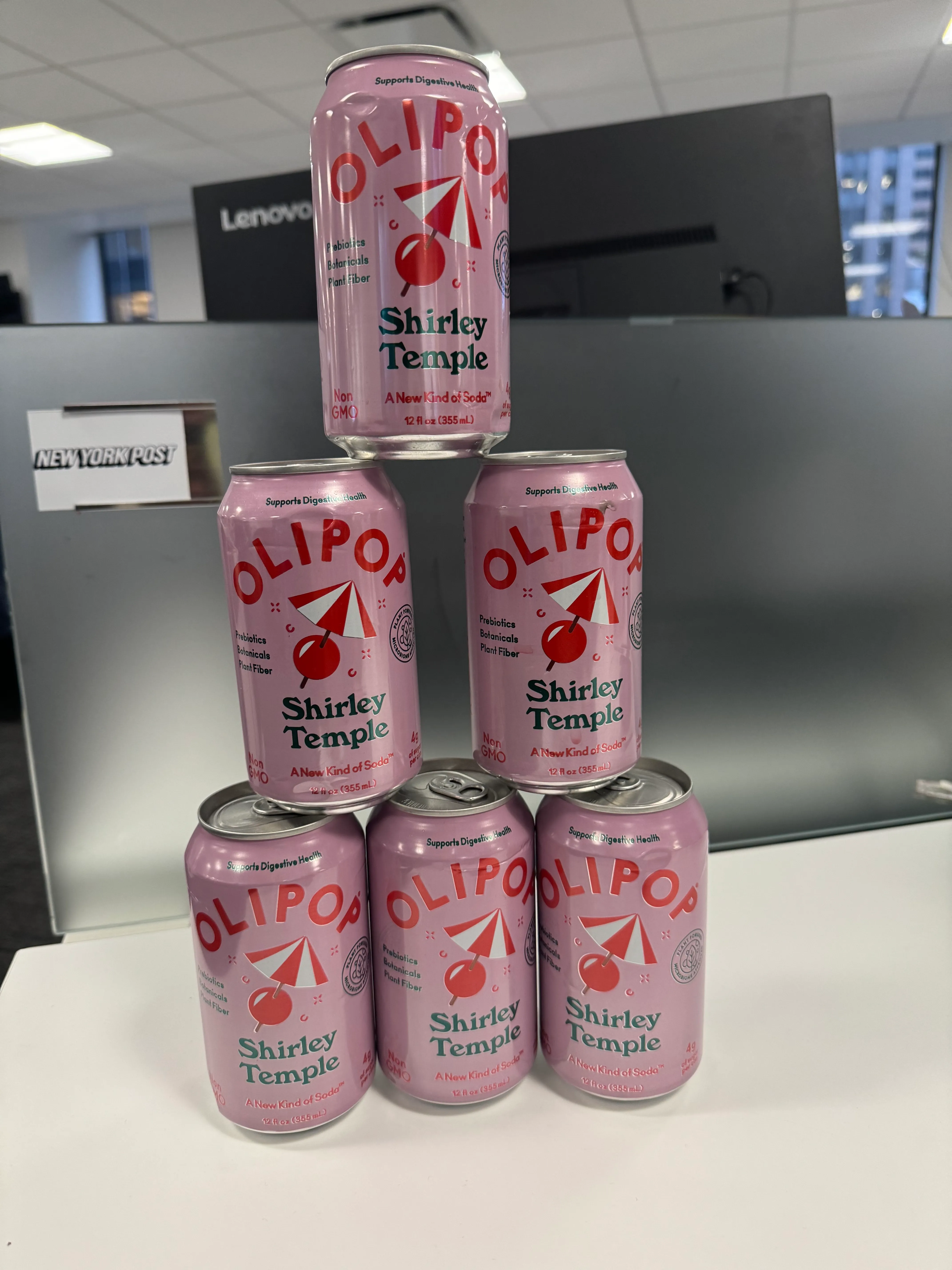 A pyramid stack of six pink OLIPOP Shirley Temple soda cans on a white desk.