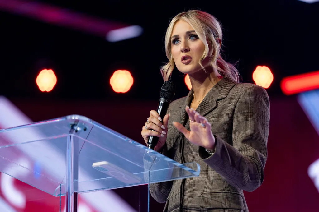A blonde woman with blue eyes speaks into a microphone at a podium, gesturing with one hand.