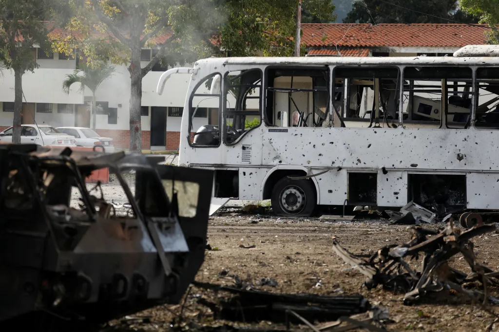 A shot-up, burned-out bus with a flat tire, next to the charred remains of a car, on a dirt road.