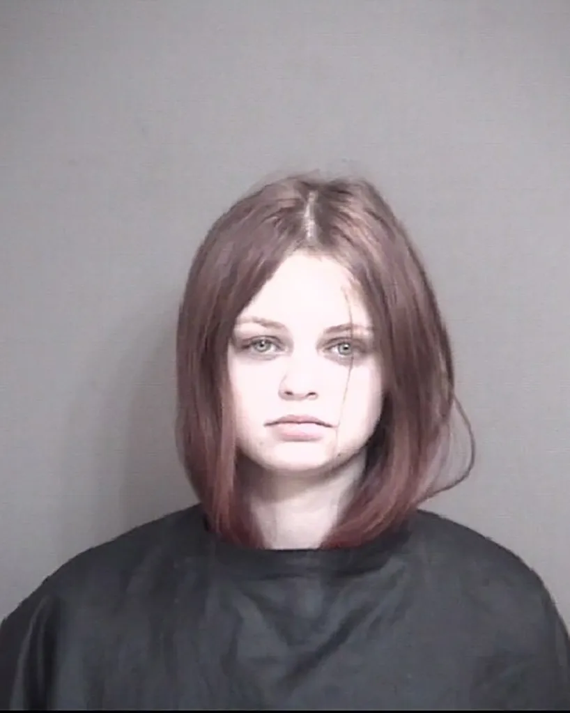 Mugshot of a young woman with shoulder-length reddish-brown hair and light eyes, wearing a dark top against a gray background.