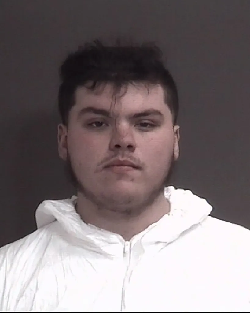 A mugshot of a young man with dark, messy hair and a faint mustache and beard, wearing a white zip-up jacket.