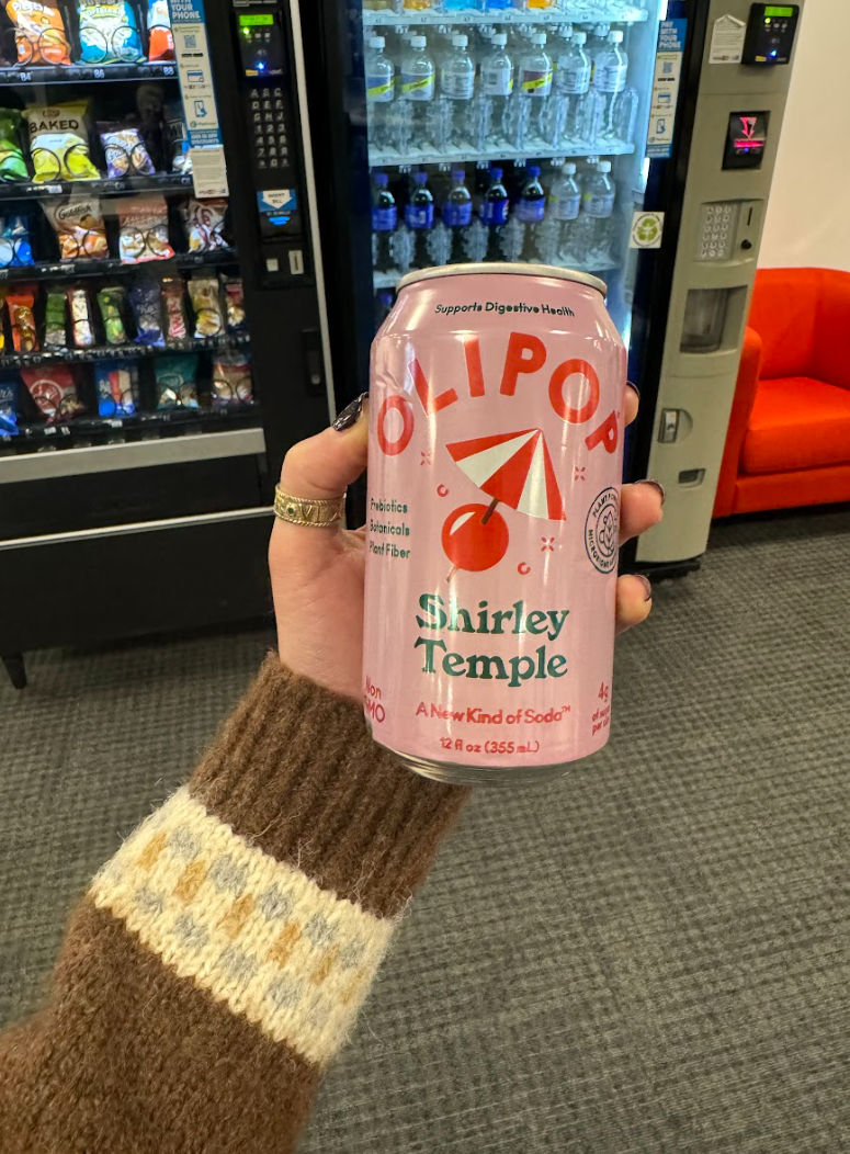 A hand holding a pink Olipop Shirley Temple soda can in front of vending machines.