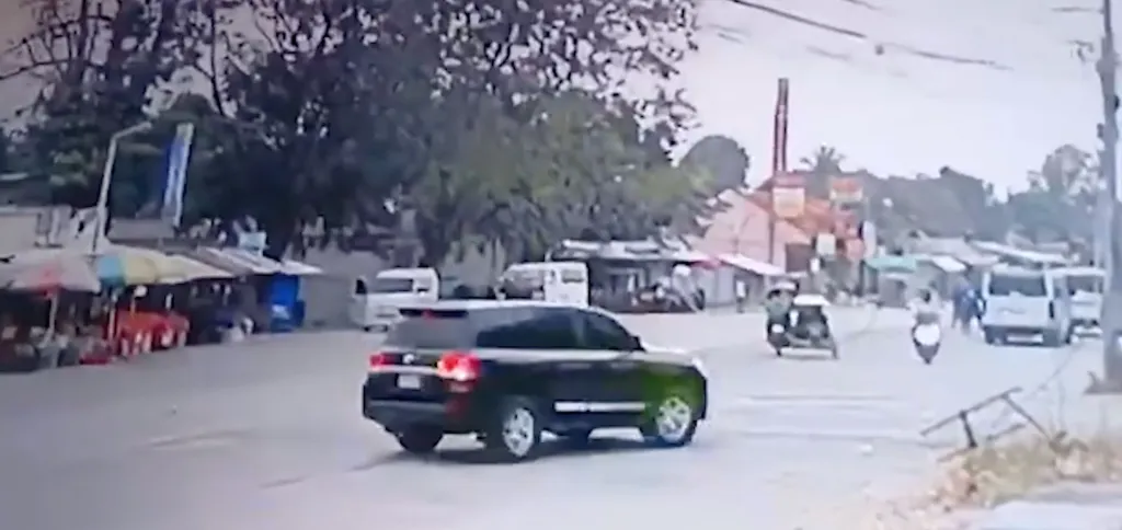 A black SUV turning left on a street with small shops, white vans, motorcycles, and trees.