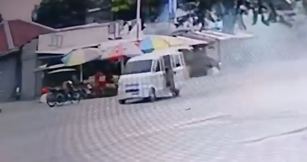 A white van with a person leaning out of the passenger side driving past parked motorcycles and an outdoor market.