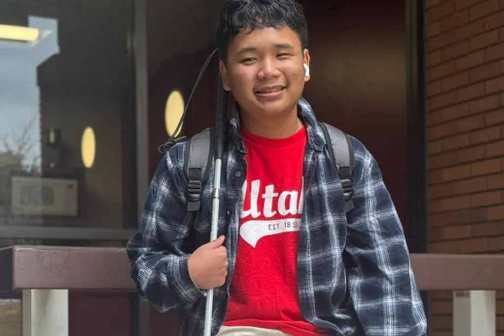 A smiling young man wearing a plaid shirt over a red Utah t-shirt, carrying a backpack and a white cane.