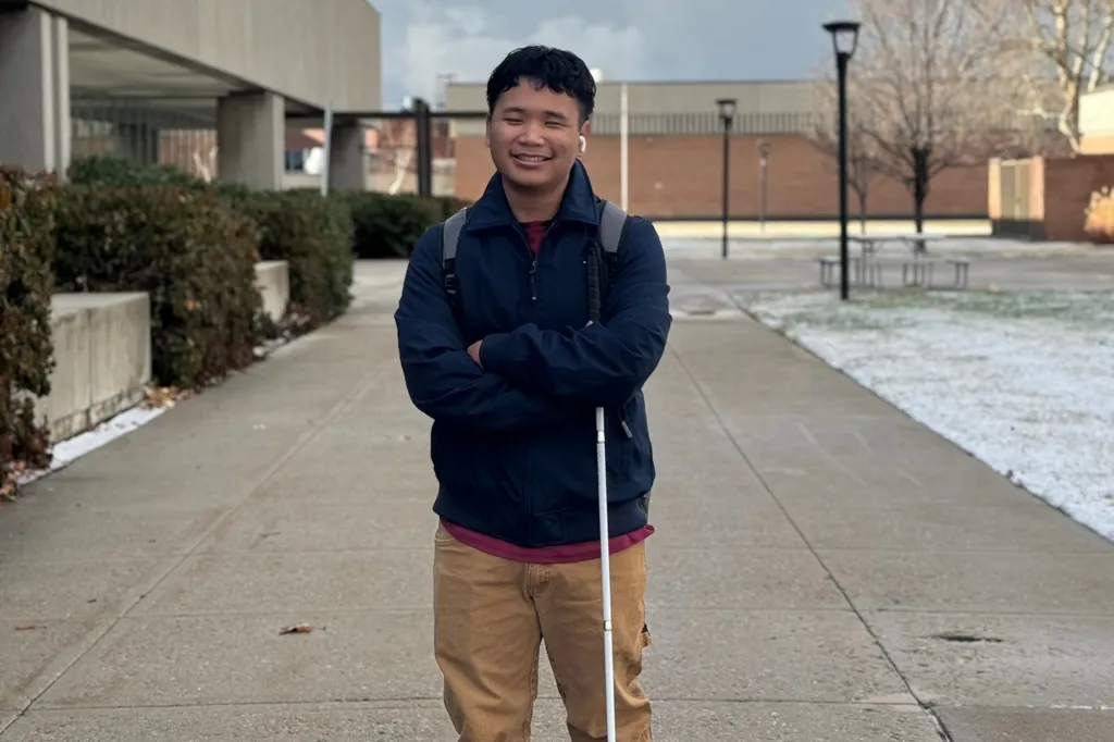 A young man with a white cane and backpack stands on a sidewalk.