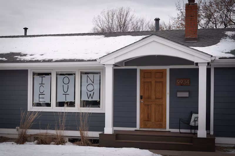 Handwritten signs reading "ICE Out Now" are displayed in the front windows of a home