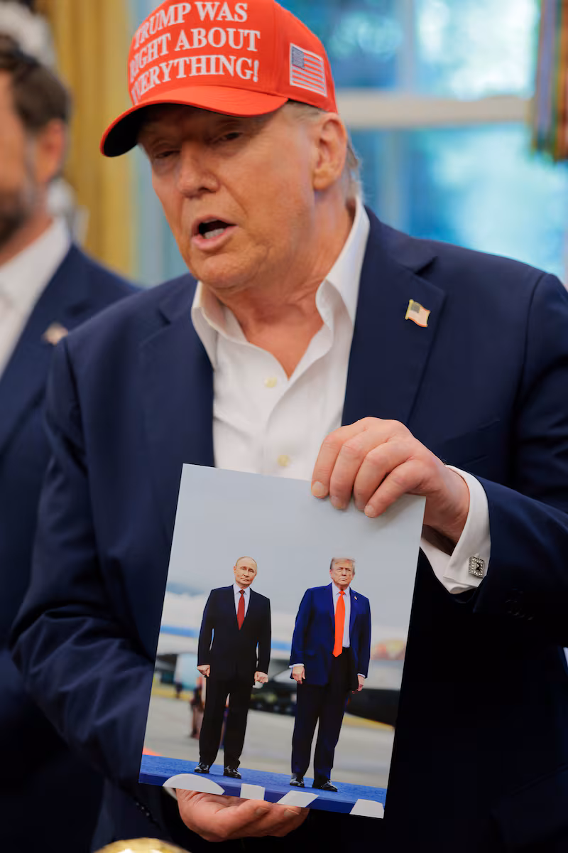 Trump in a red "Trump Was Right About Everything!" hat holds up a picture of himself with Vladimir Putin
