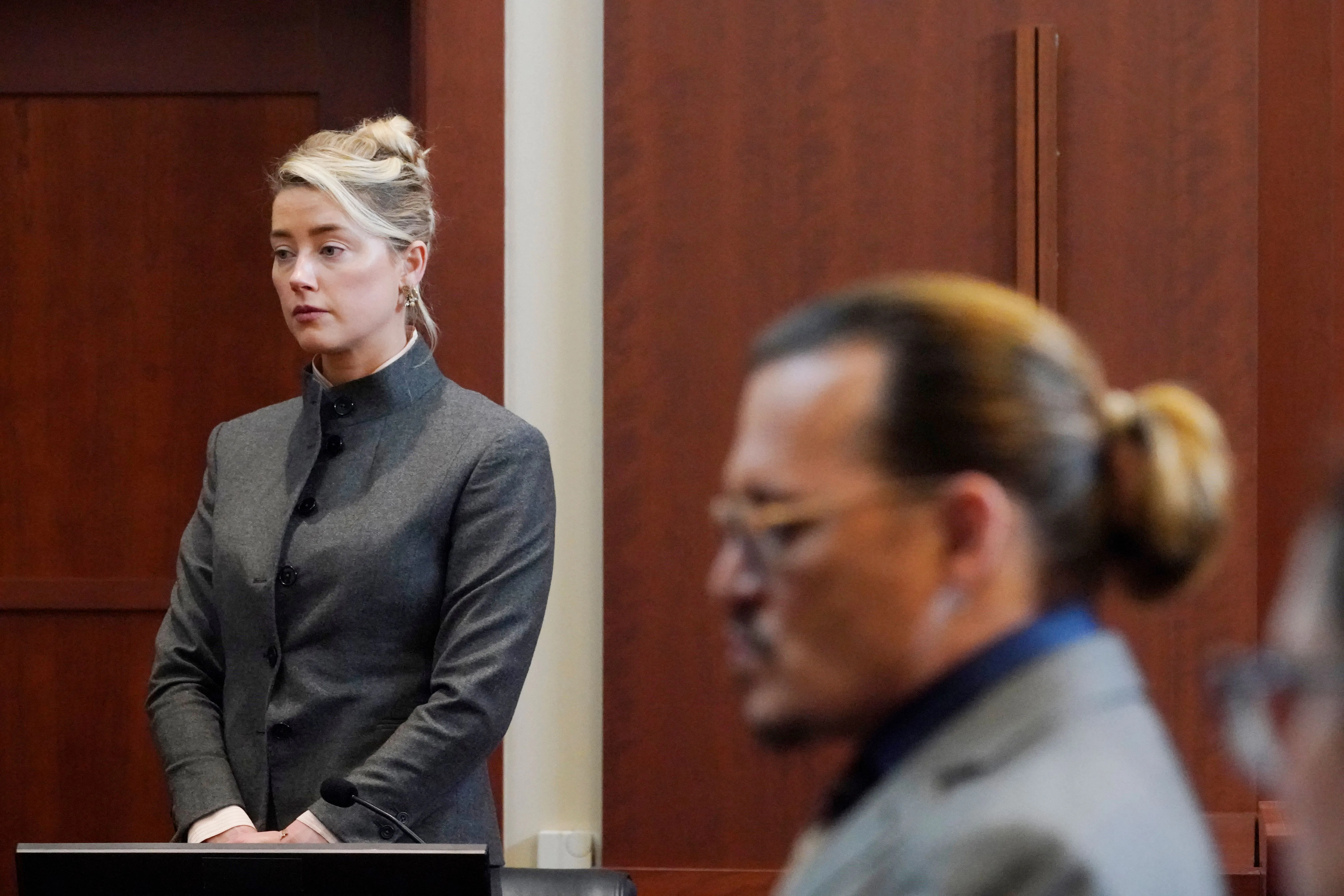 Amber Heard standing with her hands clasped in front of her, facing forward, with Johnny Depp in the foreground to the right, looking away.