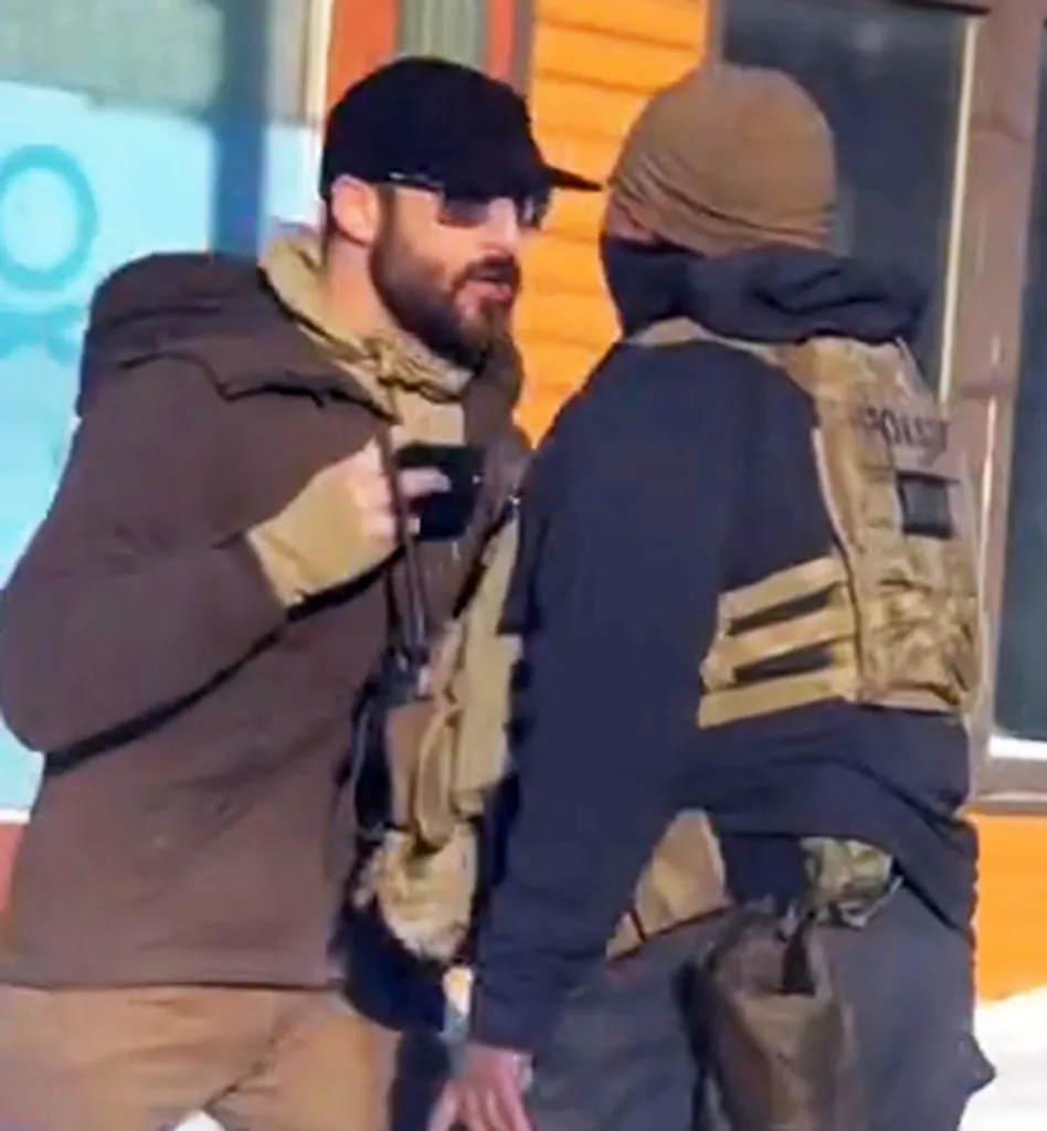Two men wearing tactical gear stand facing each other, one holding a handgun.