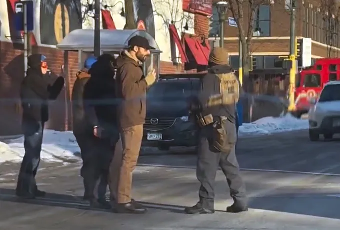 Video still of an ICE agent interacting with a group of people, one of whom is filming.