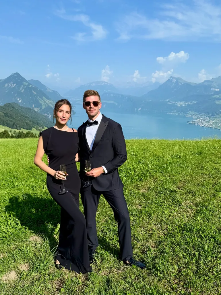 Amanda Batula and Kyle Cooke standing in a field with a lake and mountains in the background, holding champagne glasses.
