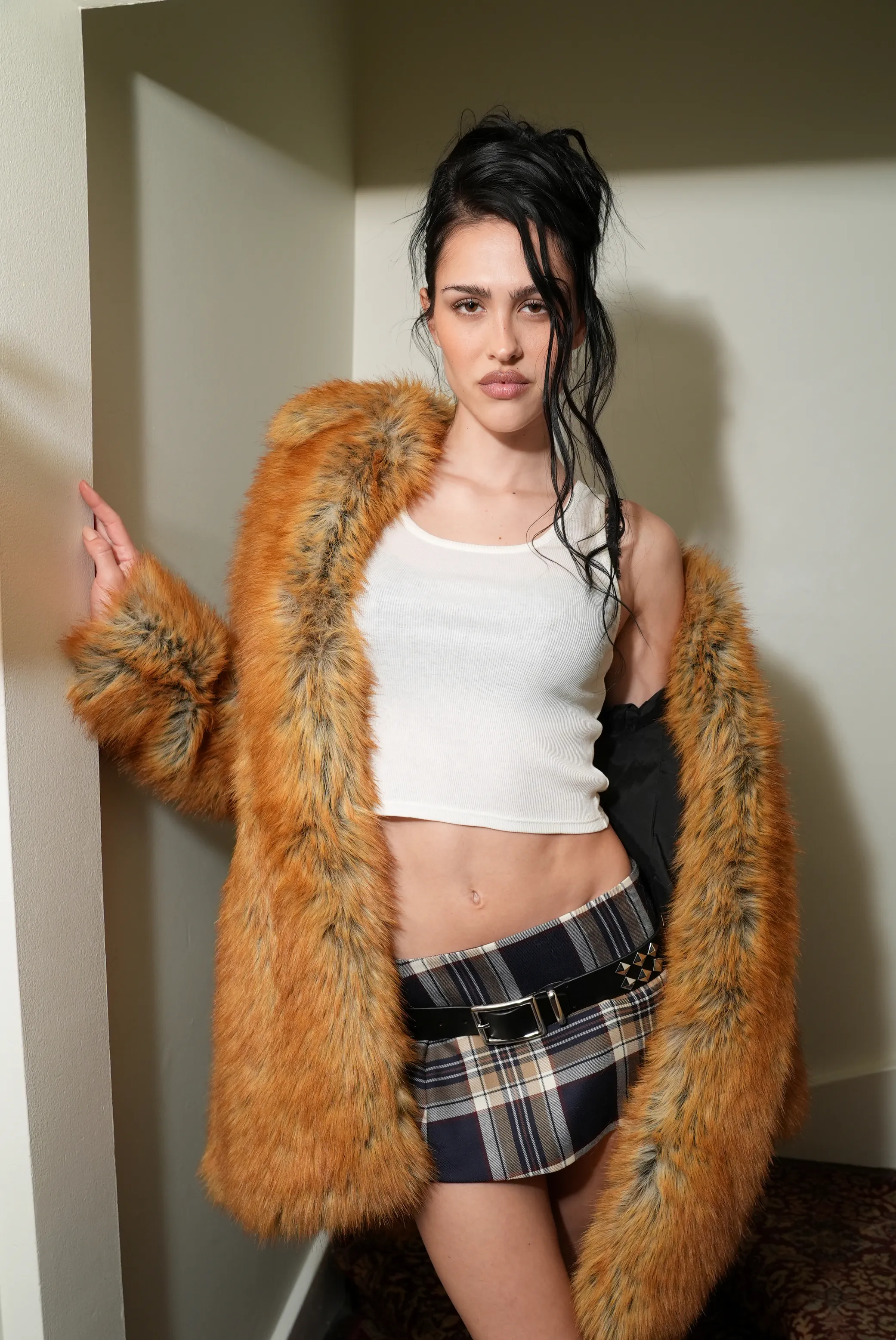 Amelia Gray Hamlin poses in a white crop top, plaid mini skirt with a studded belt, and an orange faux fur coat.