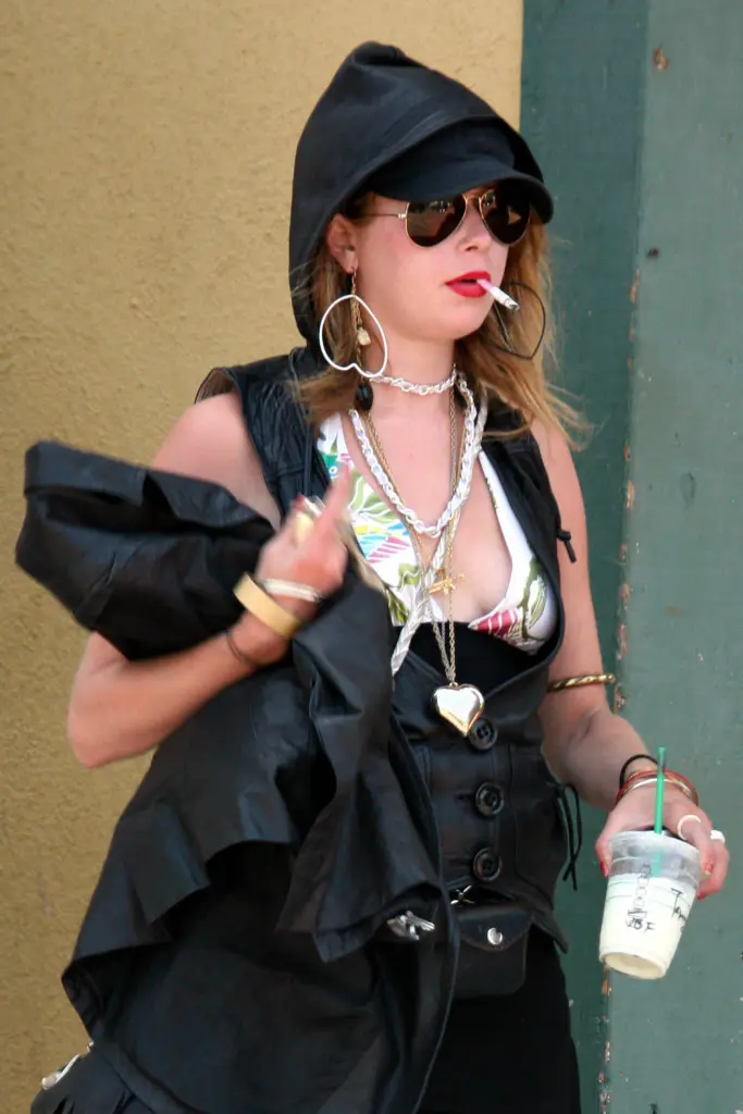 Natasha Lyonne, wearing a black hooded vest and aviator sunglasses, smokes a cigarette while holding a cold drink.