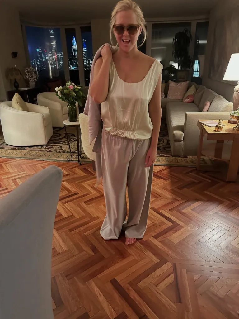 Amy Schumer in a light top and pants with a jacket over her shoulder, standing in a living room with a city night view.