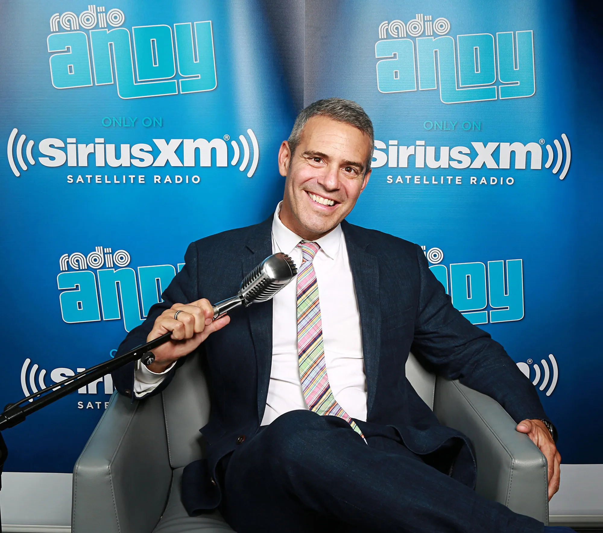 Andy Cohen sits in a grey chair, holding a microphone, in front of a blue background with